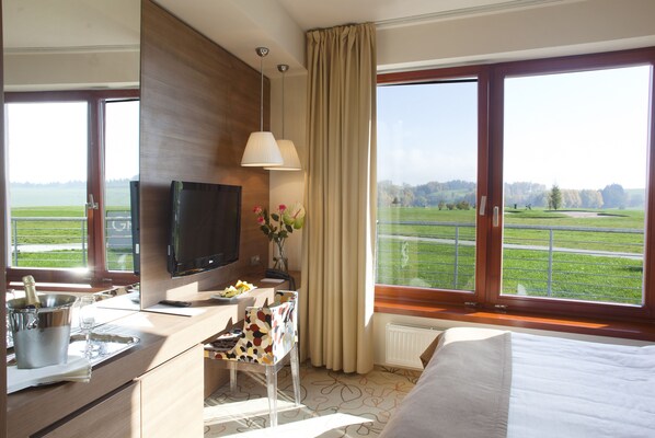 A hotel room with a bed, a desk with a TV, a window with a view of a green landscape, and a bathroom with a sink and mirror.