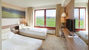 Habitación de hotel con dos camas, un ventanal con vistas a un campo verde, un televisor de pantalla plana, y un escritorio con teléfono y un jarrón de flores.