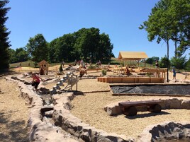 Zona de juegos al aire libre con arena, estructuras de madera y un área de descanso cubierta.