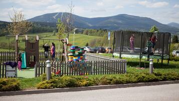A playground with swings and a climbing structure, surrounded by a wooden fence, with cars parked nearby.