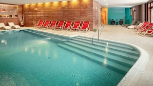 Una piscina interior con agua cristalina, rodeada de sillas reclinables rojas y un muro de madera.