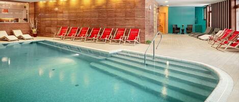 Una piscina interior con agua cristalina, rodeada de sillas reclinables rojas y un muro de madera.