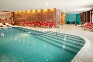 Una piscina interior con agua cristalina, rodeada de sillas reclinables rojas y un muro de madera.