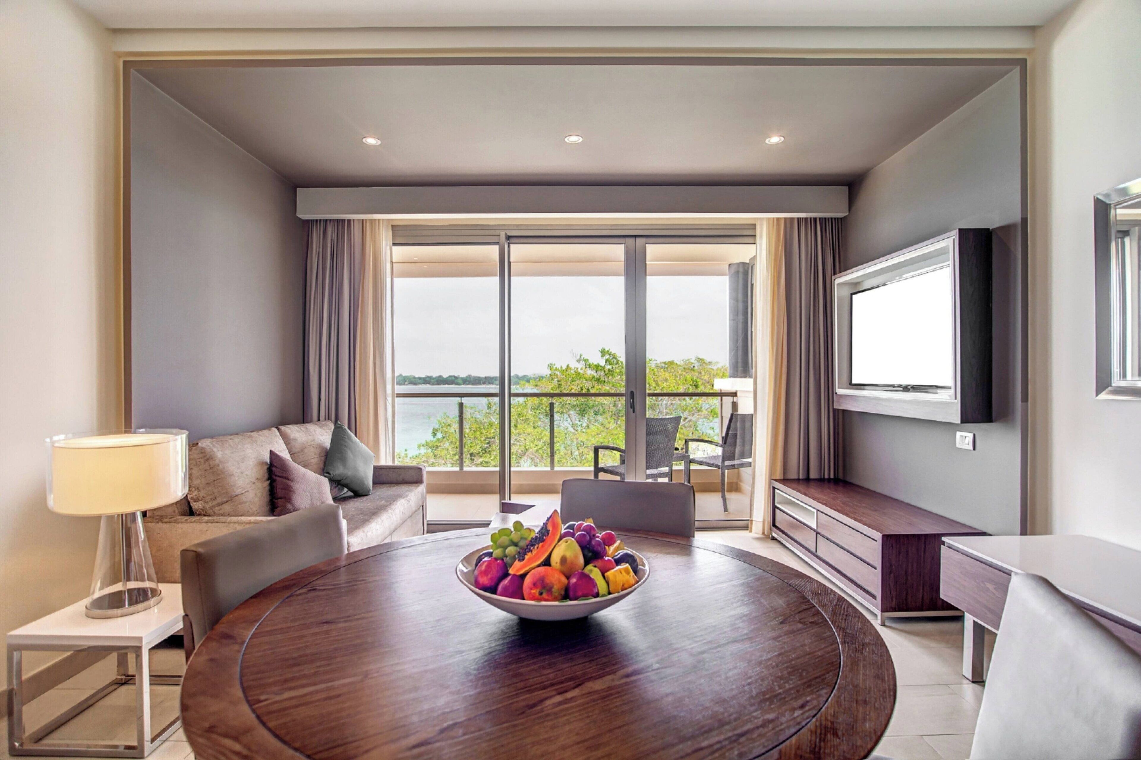 Quarto de hotel moderno com uma mesa de jantar redonda, uma tigela de frutas, um sofá e uma televisão.