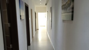 A long, narrow hallway with white walls and a tiled floor. There are several doors on the left side, and artwork is displayed on the walls. - Malta Rent Rooms (Gzira)