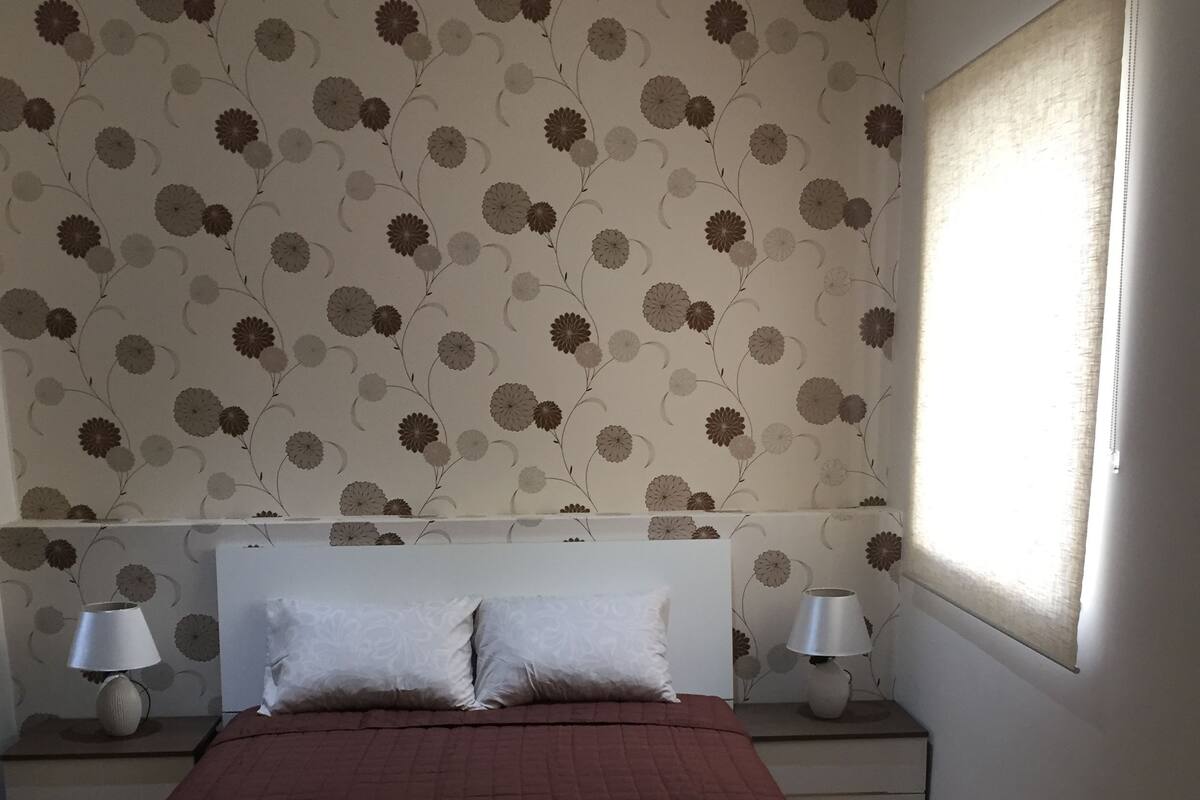 A bedroom with a large bed, two bedside tables with lamps, a dresser, and a window with curtains.