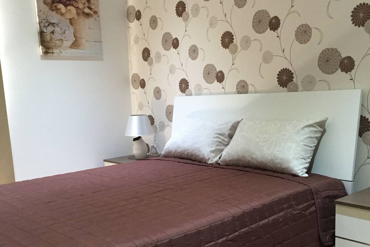 A bedroom with a bed, a nightstand, a lamp, and a wall-mounted air conditioner.
