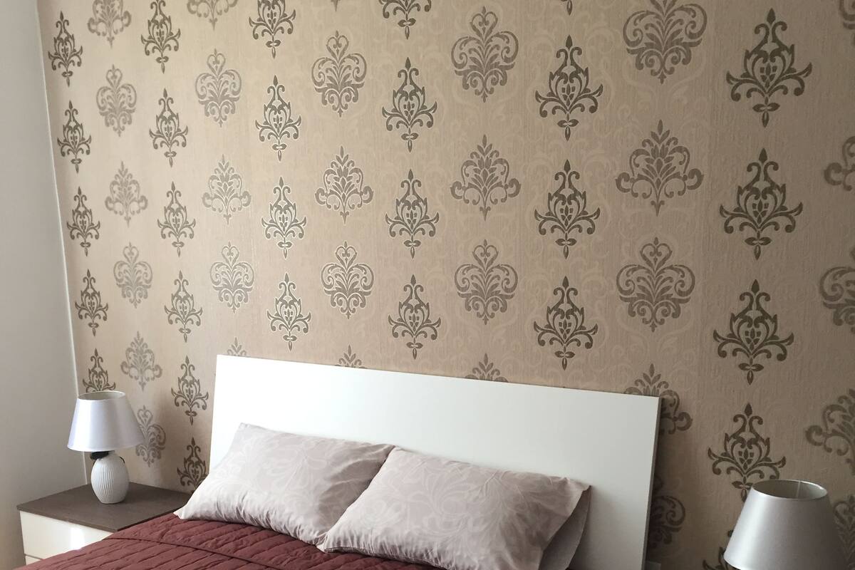A neatly made bed with a red quilt, two bedside lamps, and a patterned wallpaper.
