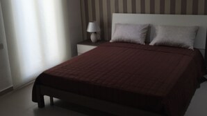 A bedroom with a large bed, a nightstand, a lamp, and a window with blinds. - Malta Rent Rooms (Gzira)