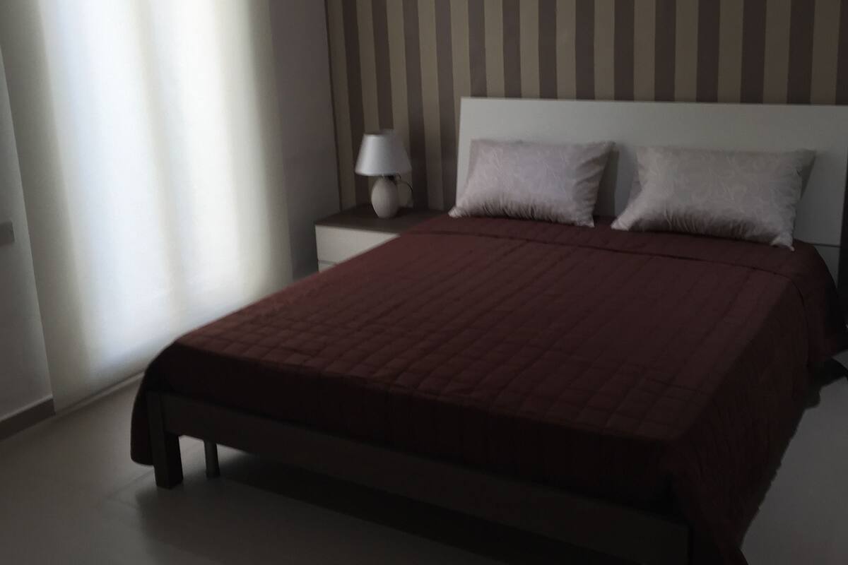 A bedroom with a large bed, a nightstand, a lamp, and a window with blinds.