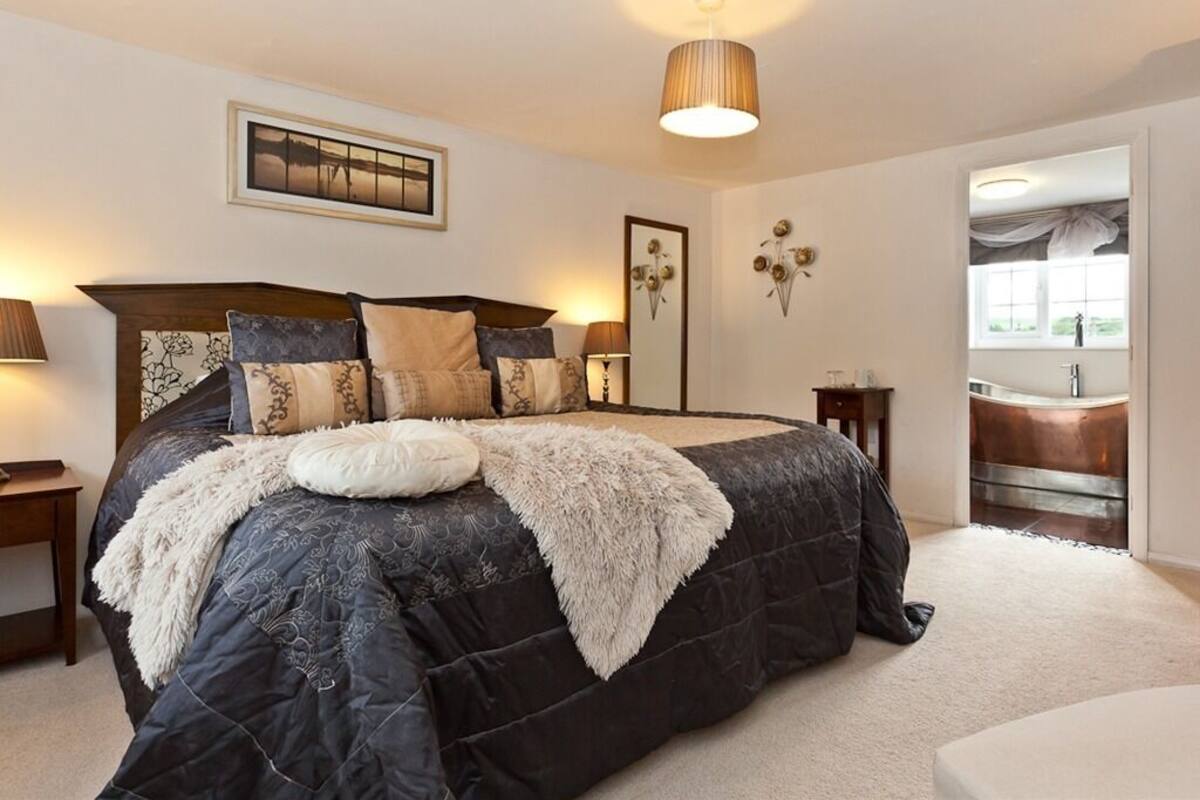 Une chambre avec un grand lit, une table de chevet, une lampe, un miroir fixé au mur et une vue sur la salle de bain par une porte ouverte.