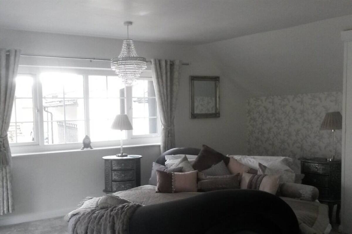 Une chambre à coucher comprenant un lit, une table de chevet, une commode, un lustre et une fenêtre avec des rideaux.