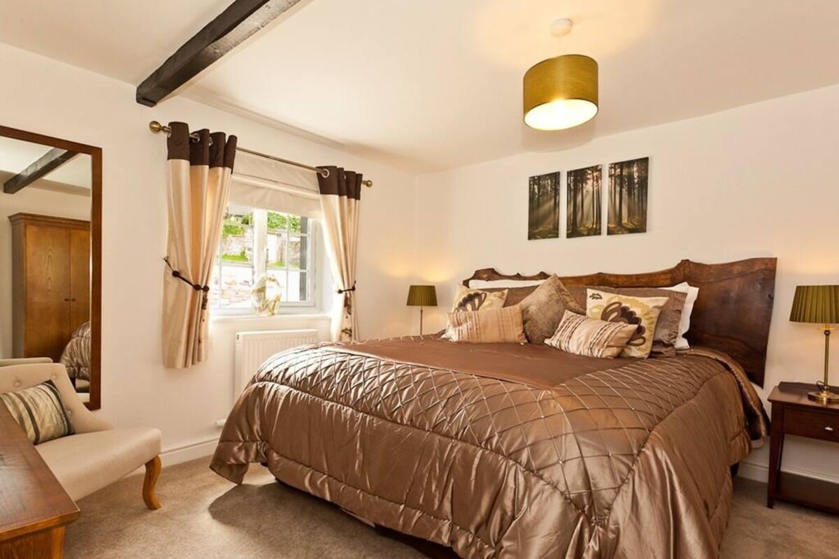 Une chambre à coucher avec un grand lit, une tête de lit en bois, une table de chevet, une lampe, une fenêtre avec des rideaux et un miroir.