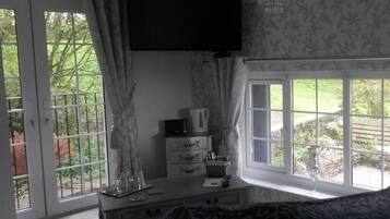 A room with a television mounted on the wall, a desk with a lamp, and a window with a view of greenery outside.