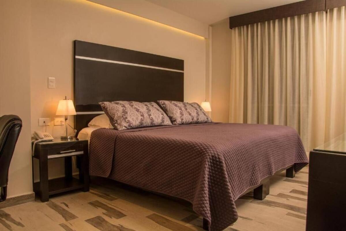 Ein Hotelzimmer mit einem großen Bett, zwei Nachttischen mit Lampen, einem Telefon und Blick durch Vorhänge nach draußen.