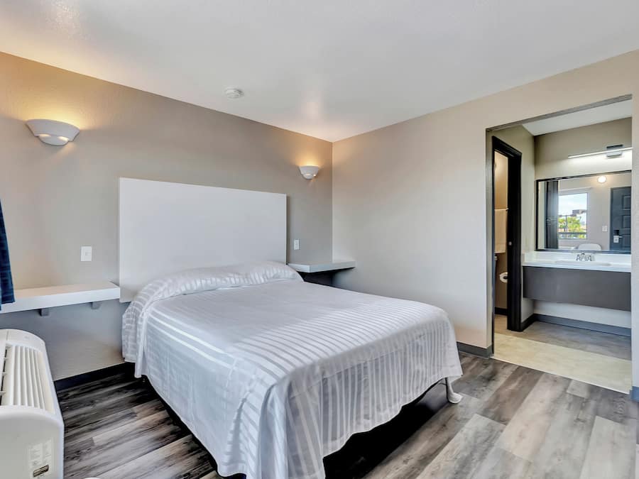 A hotel room with a bed, bedside tables, a wall-mounted light, a ceiling vent, and a bathroom visible through an open door.