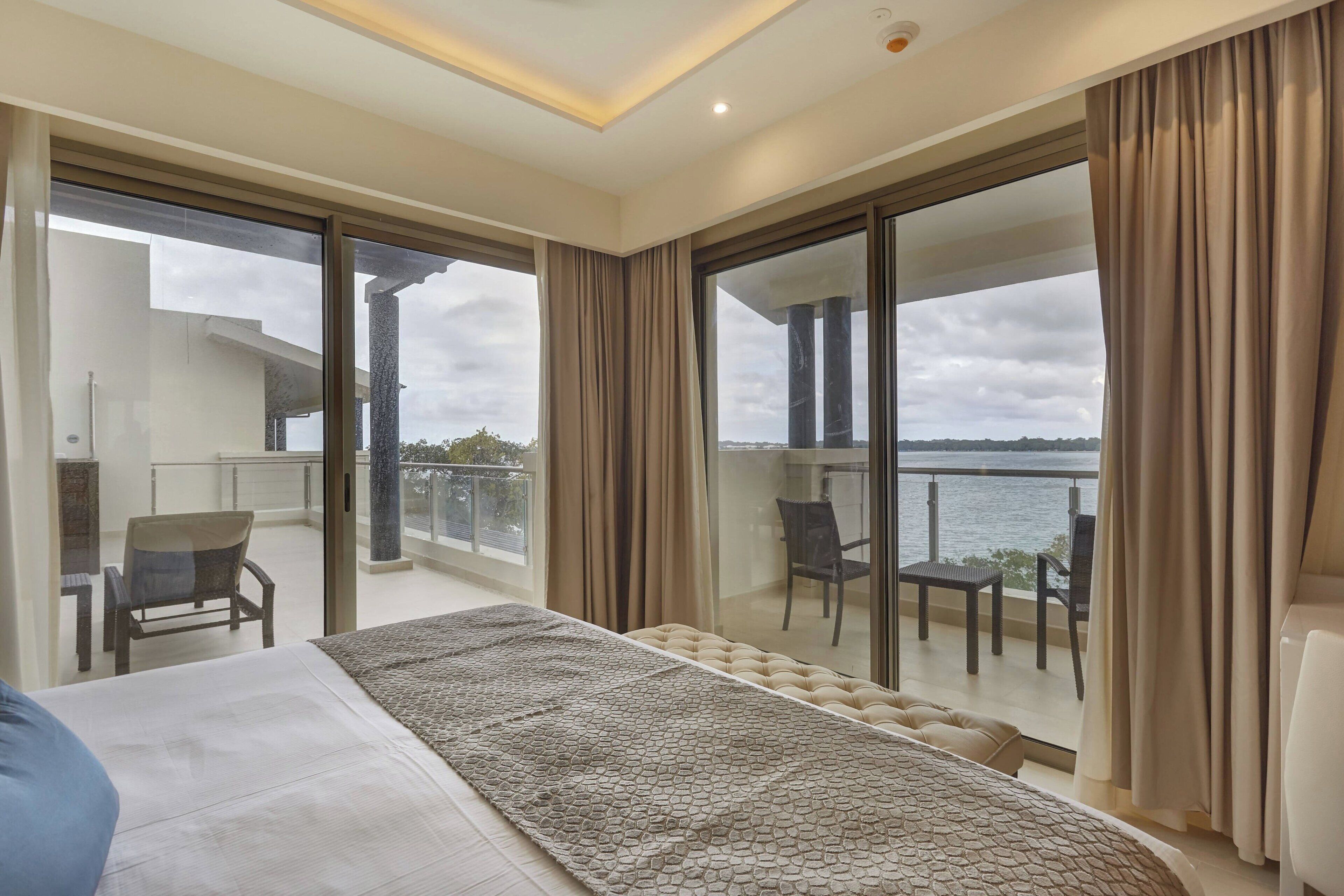 A modern bedroom with a large bed, a balcony with a view of the water, and a separate seating area with chairs.