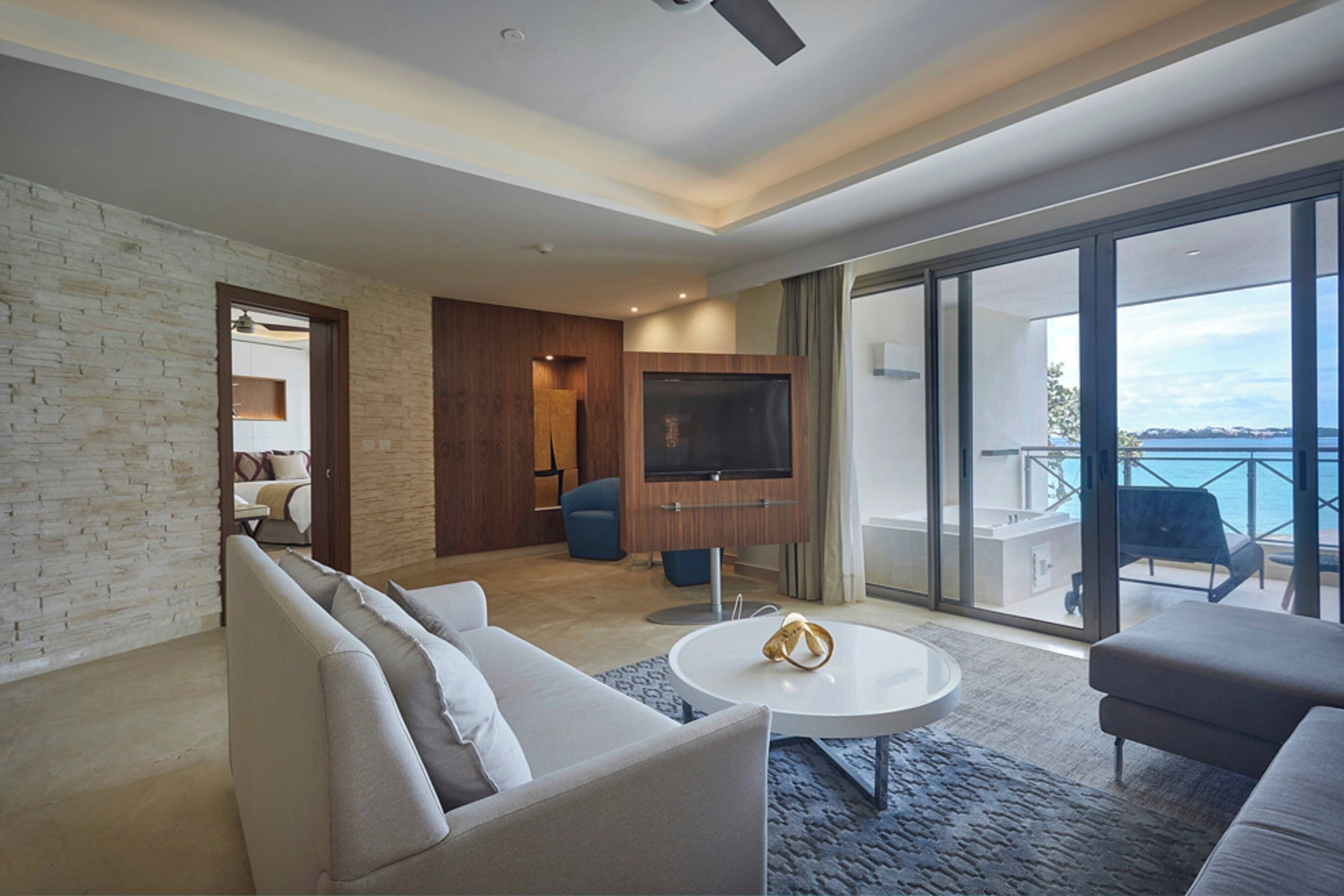 Una sala de estar moderna con un sofá, una mesa de centro y un televisor. Hay un balcón con vista al mar.