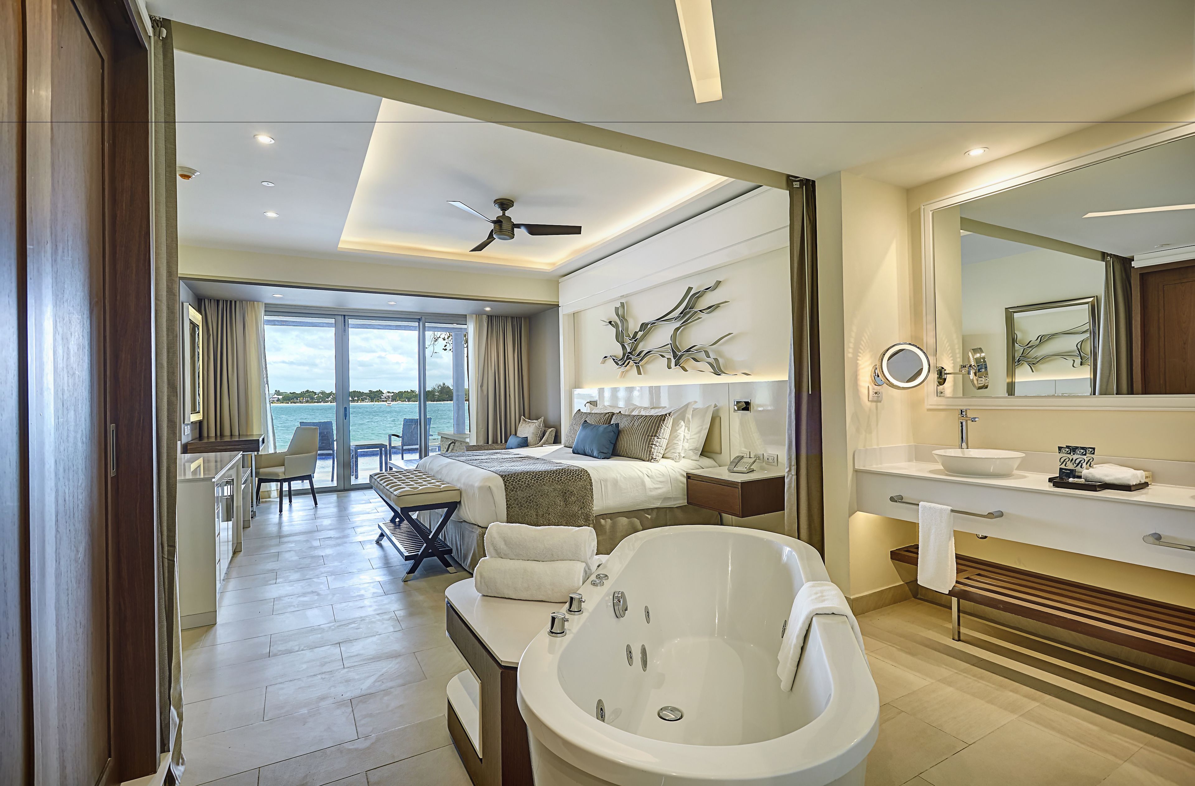 A modern hotel room with a large bathtub, a bed with a view, a dining area, and a bathroom with a large mirror and sink.