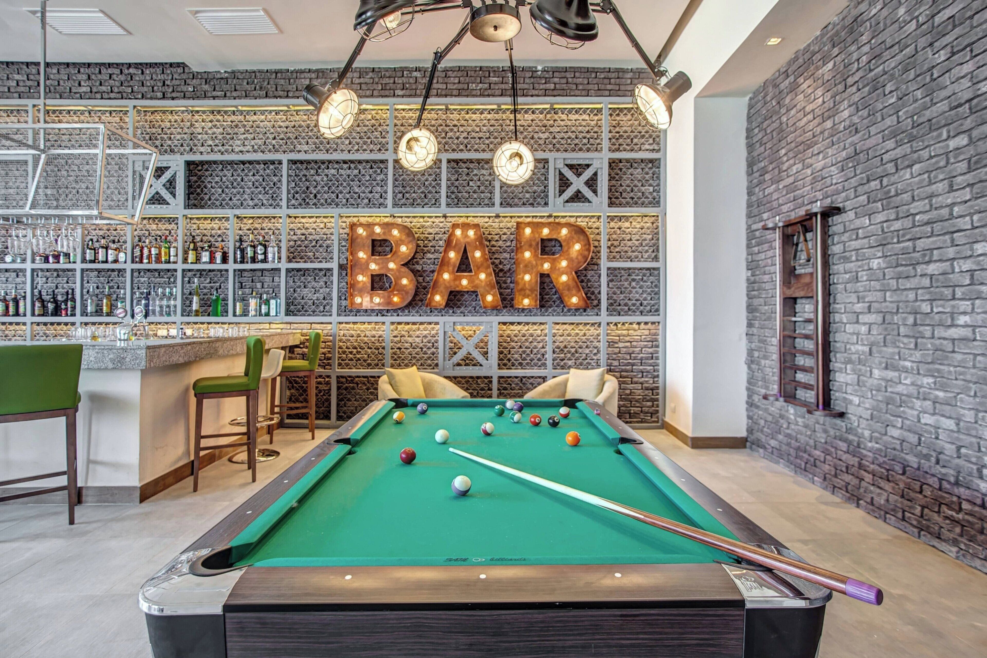 A pool table with a bar area in the background, featuring a 'BAR' sign and shelves of bottles.