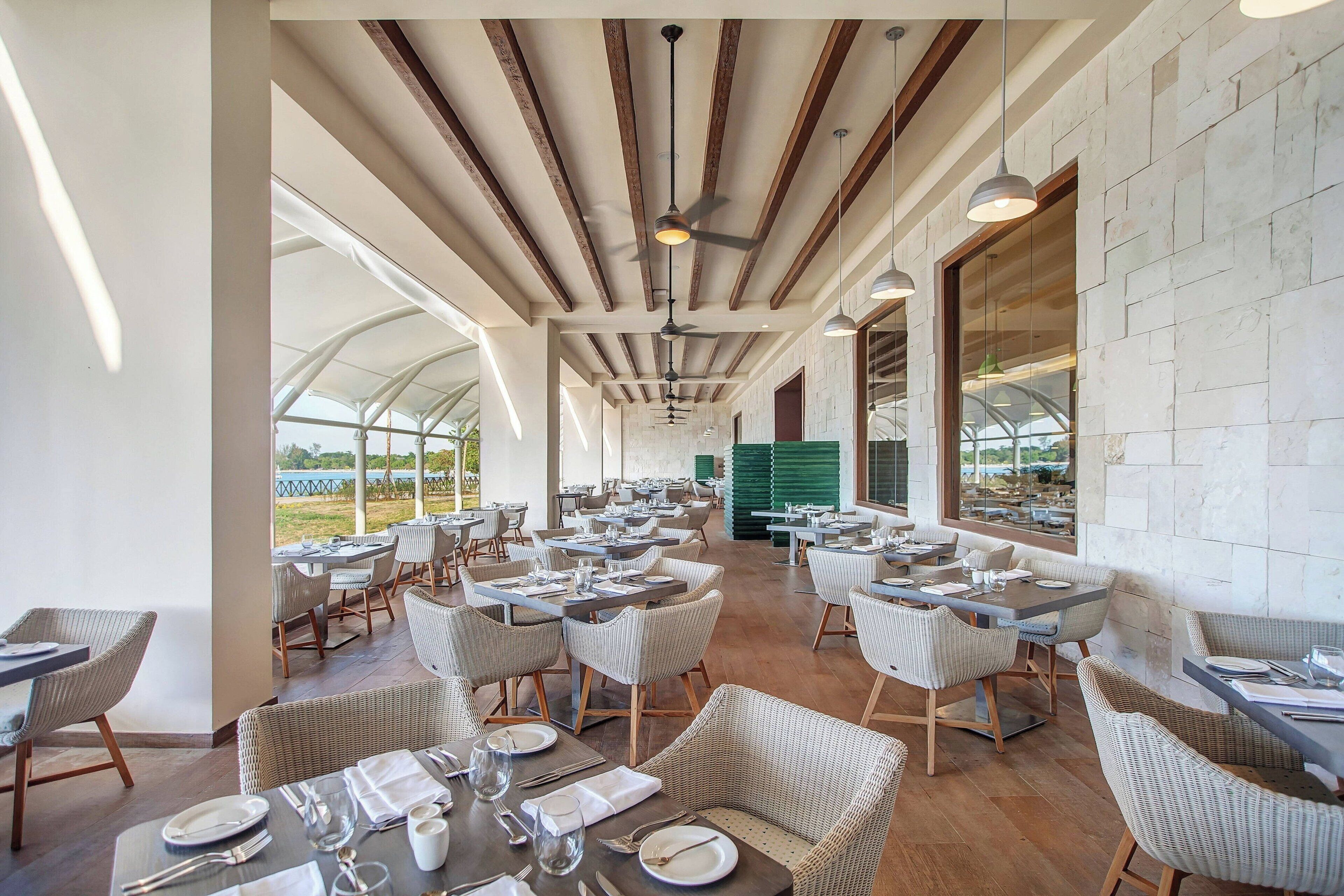 A spacious dining area with high ceilings, wooden beams, and large windows offering views of the outdoors. Tables are set with white tablecloths and wicker chairs.