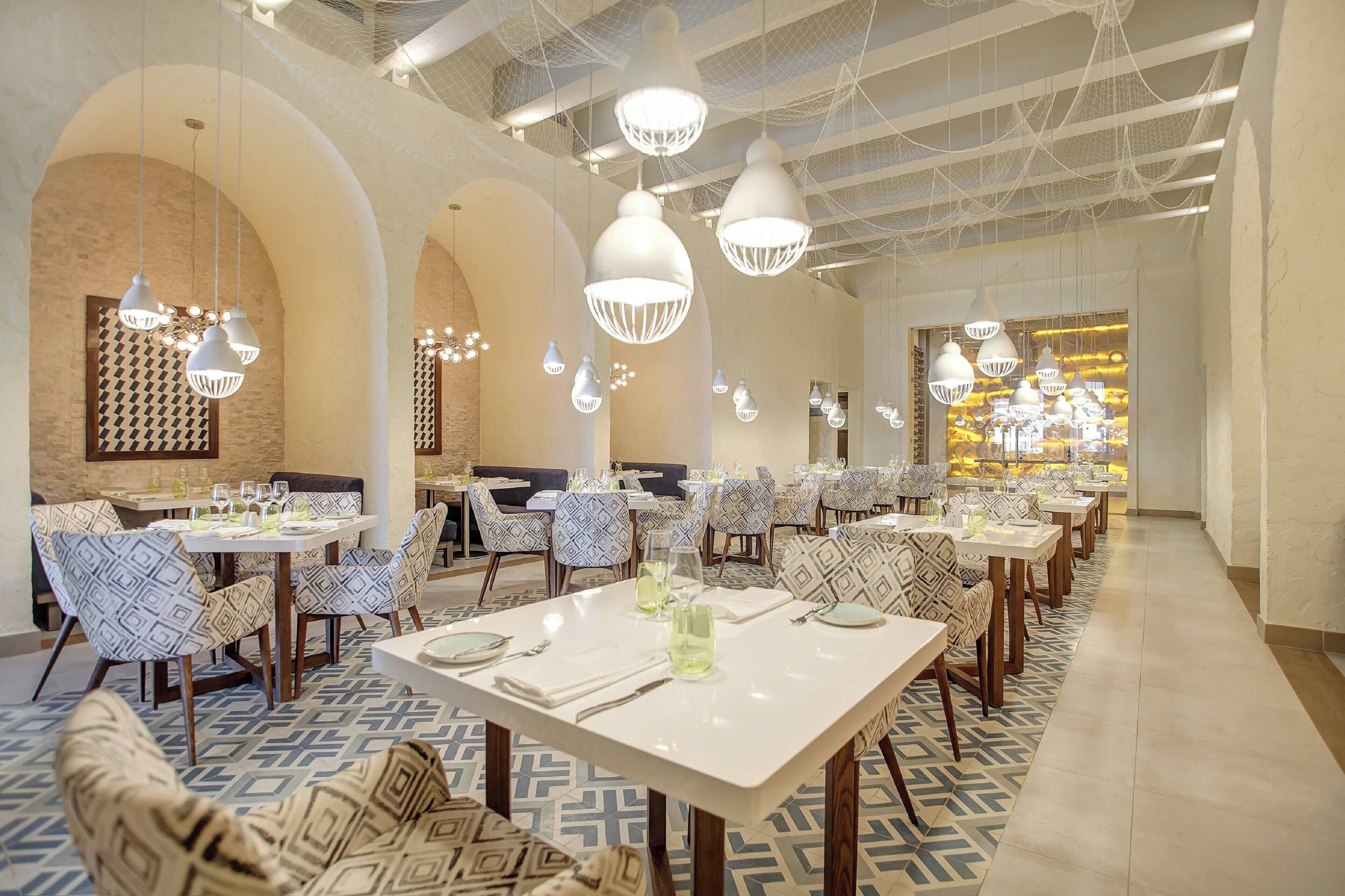 A restaurant with multiple dining tables and chairs, hanging pendant lights, and a patterned floor.