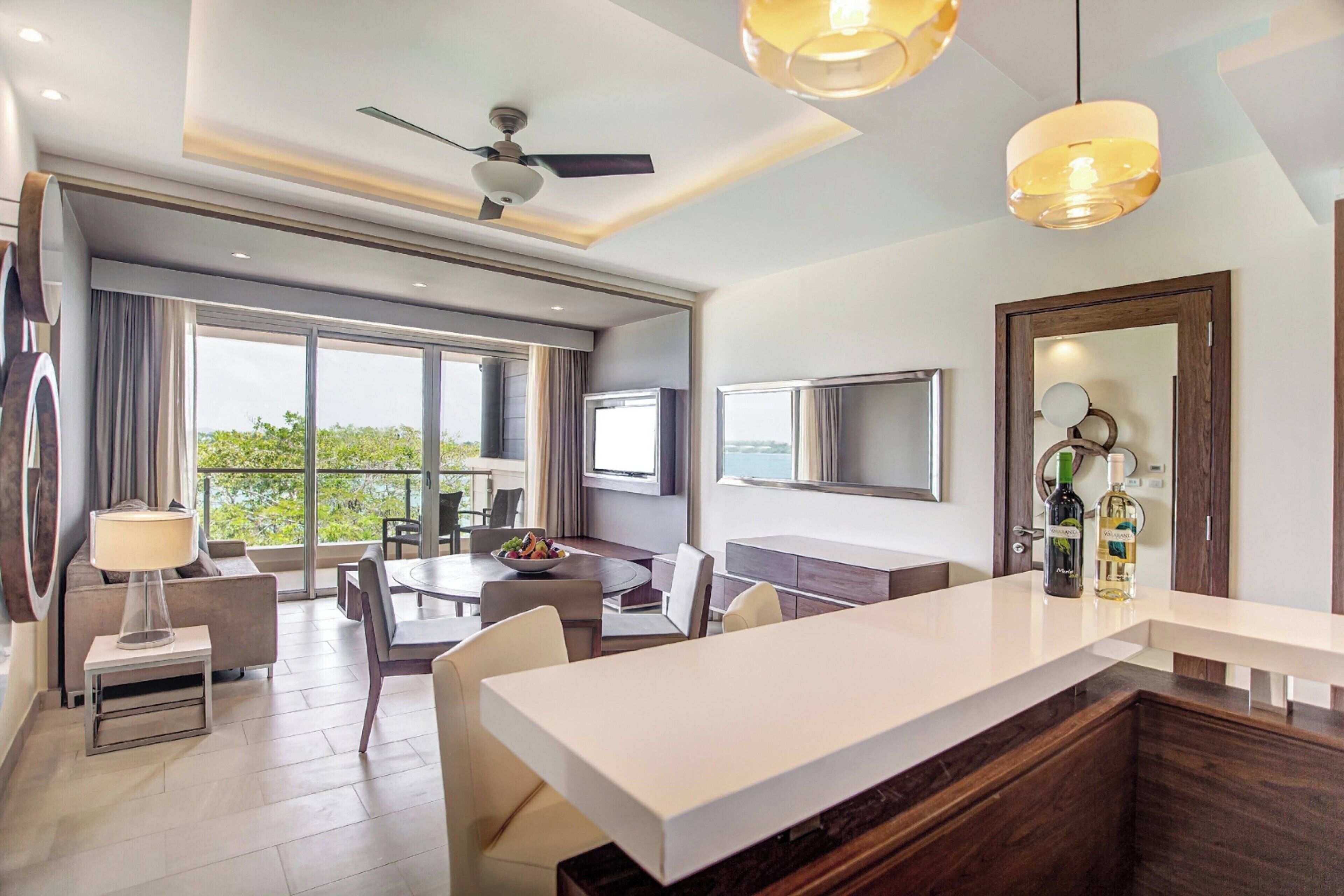A modern hotel room with a large balcony, a dining area, a kitchen island, and a flat-screen TV.