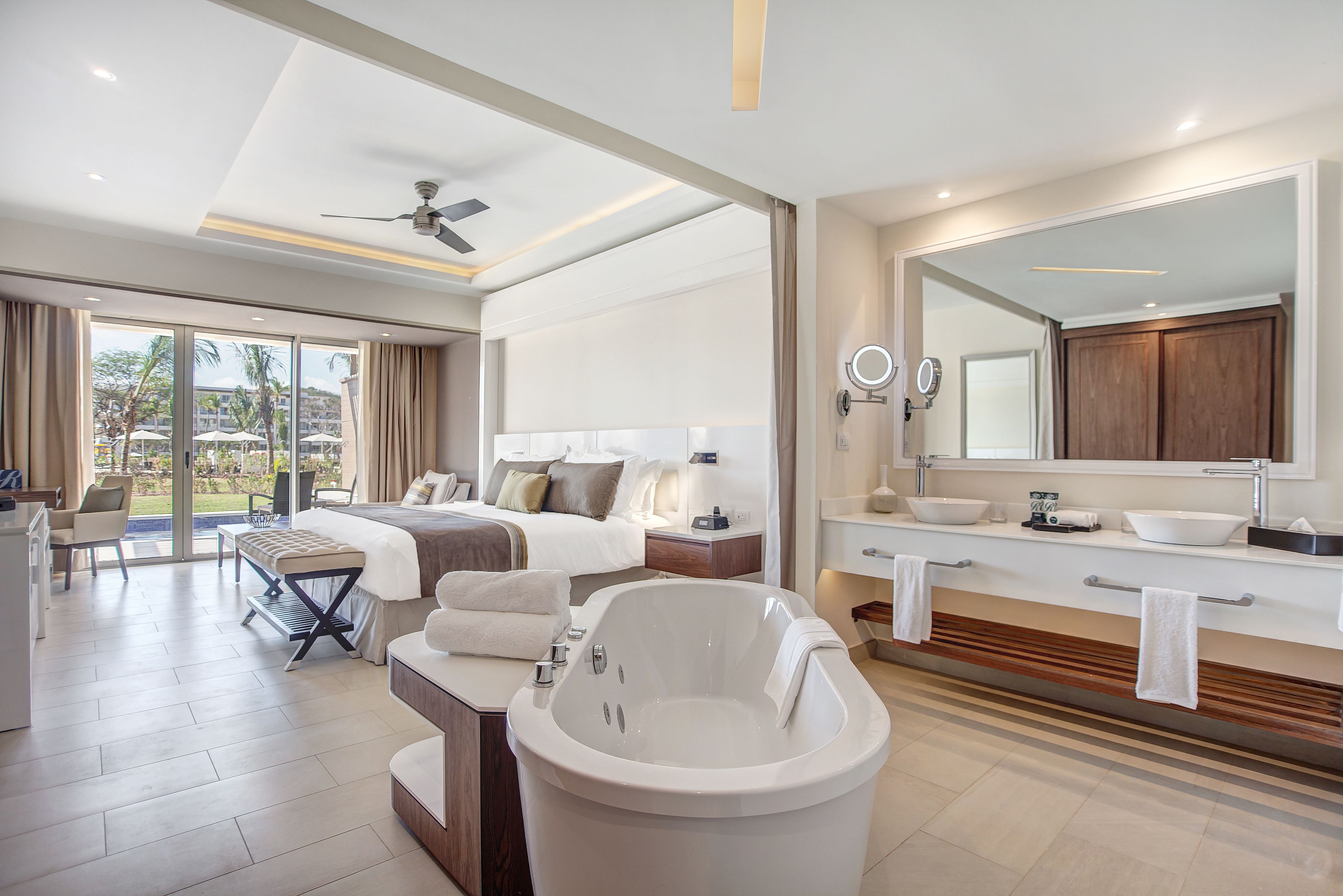 A modern hotel room with a large bed, a bathtub, a vanity with a mirror, and a balcony with a view of palm trees.