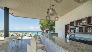 A bar area with a view of the beach, outdoor seating, and a bar counter with shelves of bottles. - Royalton Negril, An Autograph Collection All-Inclusive Resort (Negril)