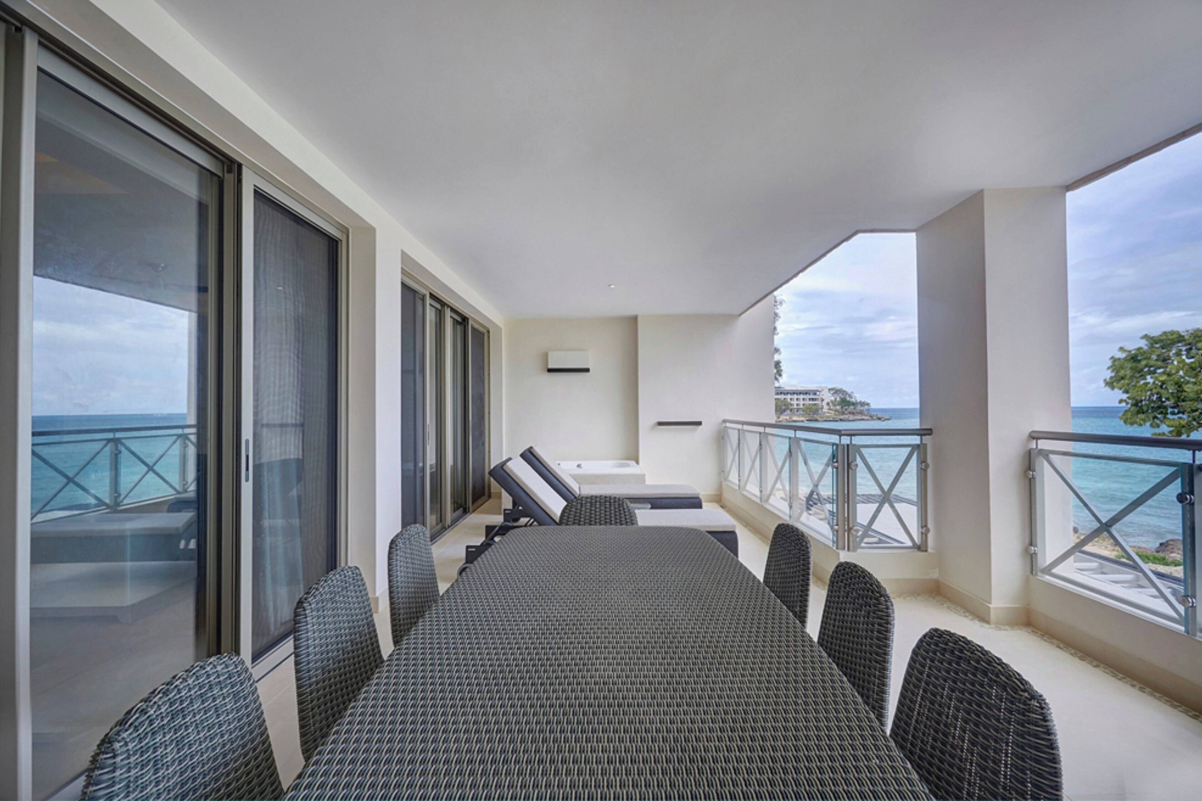 A balcony with a dining table and chairs, large windows, and a view of the sea.