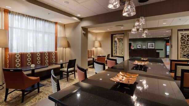 A hotel lobby with a long reception desk, seating area with patterned cushions, and modern pendant lighting. - Hampton Inn Morristown, I-81 (Morristown)