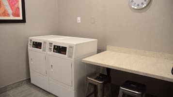 A laundry room with two stacked washing machines, a small stool, and a wall-mounted clock.