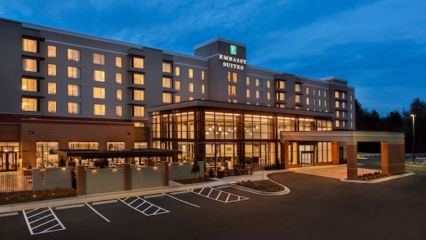 Un hotel moderno con un llamativo letrero de "Embassy Suites", un amplio espacio de esparcimiento al aire libre y una entrada bien iluminada.
