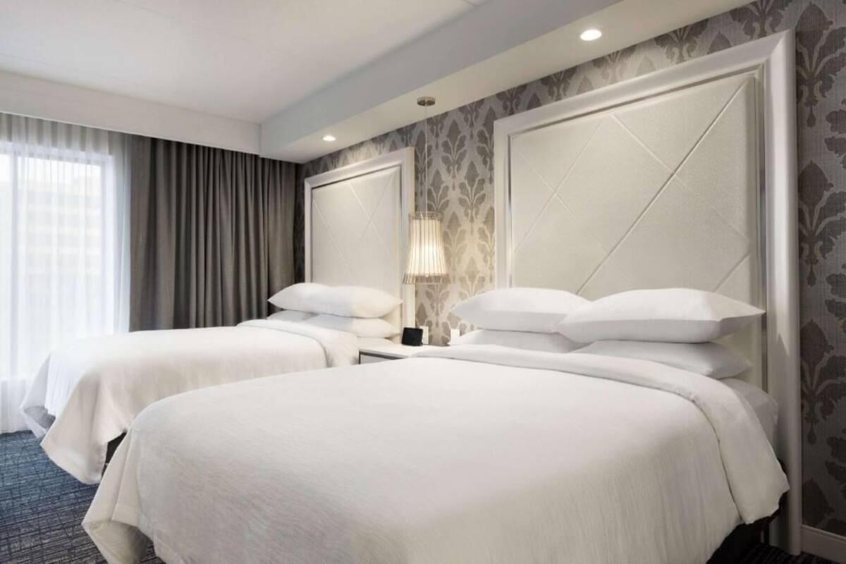 A hotel room with two beds, a nightstand, a lamp, and a window with curtains.