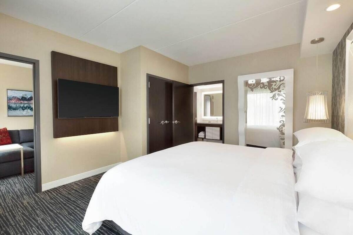 Une chambre d’hôtel dotée d’un grand lit, d’un téléviseur à écran plat fixé au mur, d’une armoire, d’une salle de bain avec un miroir et d’un petit coin salon avec un canapé et une table.