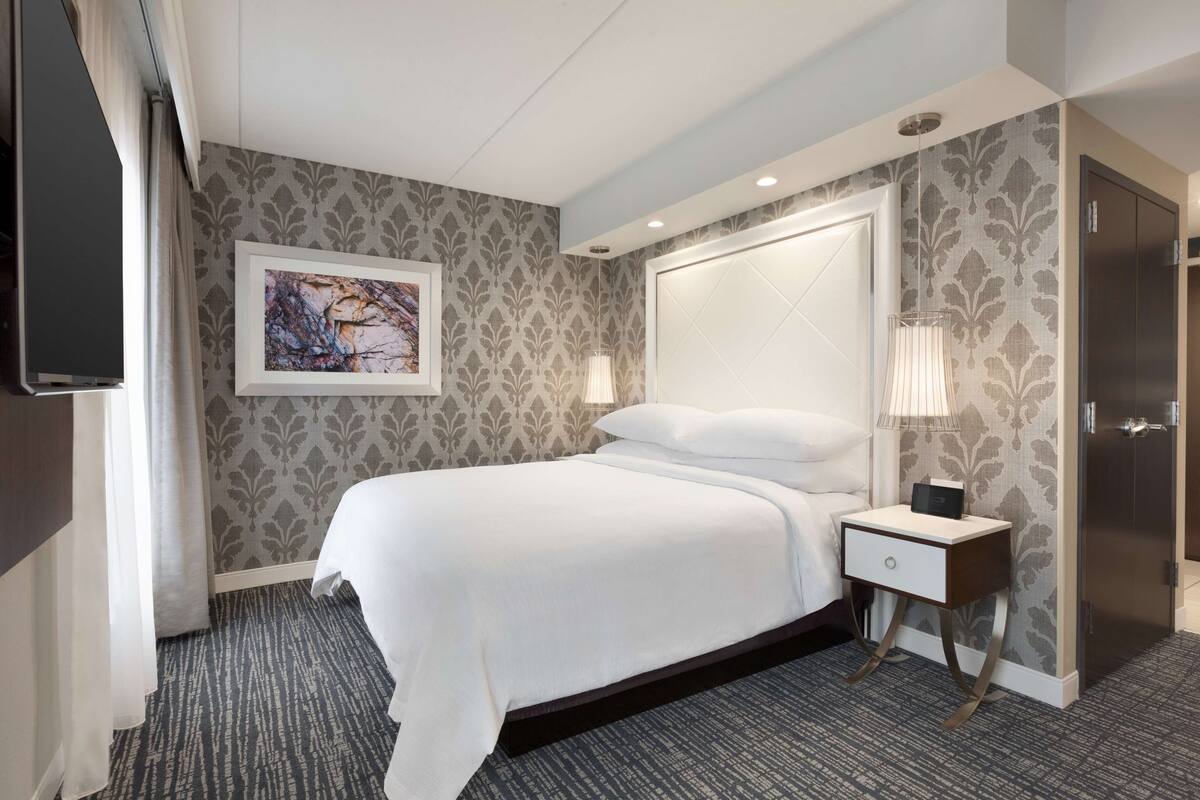 A hotel room with a bed, bedside table, lamp, artwork, and a wardrobe.