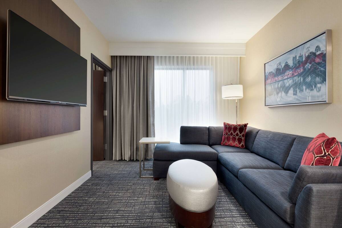 A hotel room with a large flat-screen TV, a grey sofa, a small ottoman, a side table, and a cityscape painting on the wall.