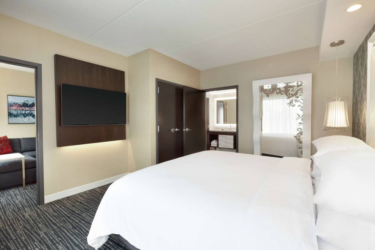 A hotel room with a large bed, a flat-screen TV mounted on the wall, a dark wooden wardrobe, a bathroom with a mirror, and a window with curtains.