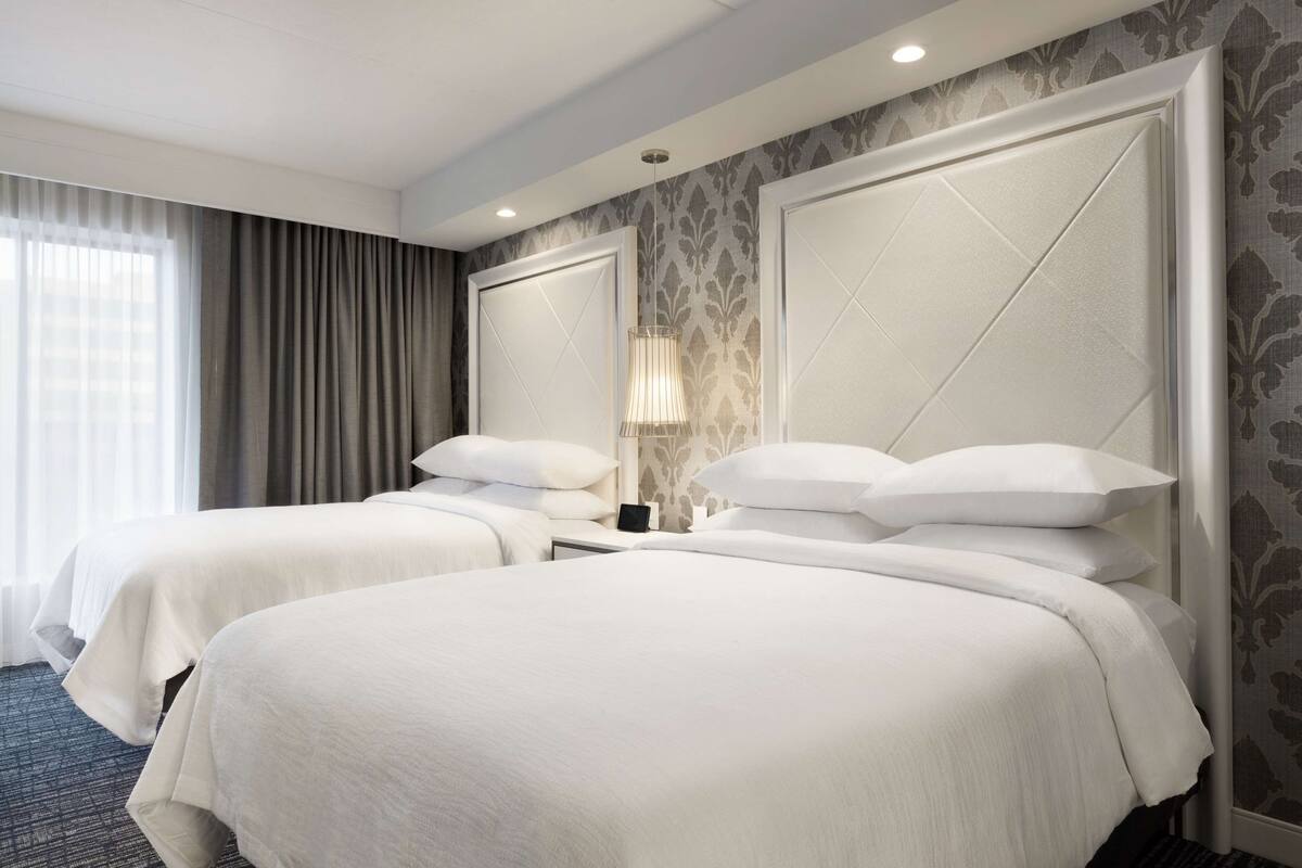 A hotel room with two beds, a nightstand, a lamp, and a large window with curtains.