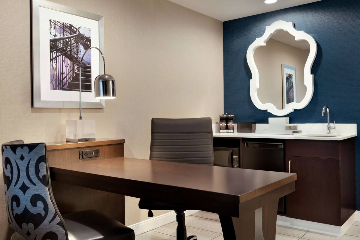 A modern hotel room with a wooden dining table, a black upholstered chair, a desk with a lamp, a small kitchenette, and a large mirror on the wall.