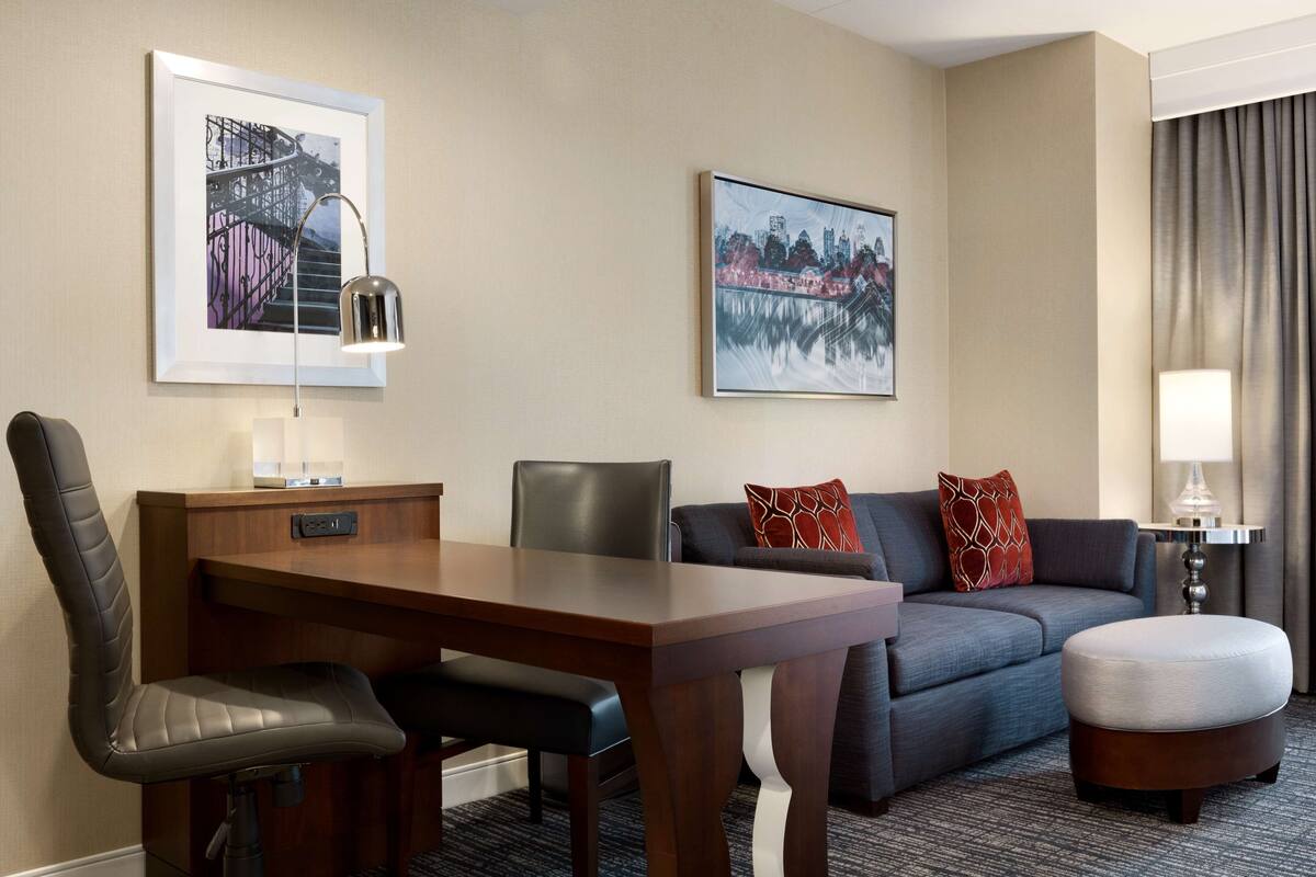 A hotel room with a dining table, a sofa, an ottoman, and two framed pictures on the wall.