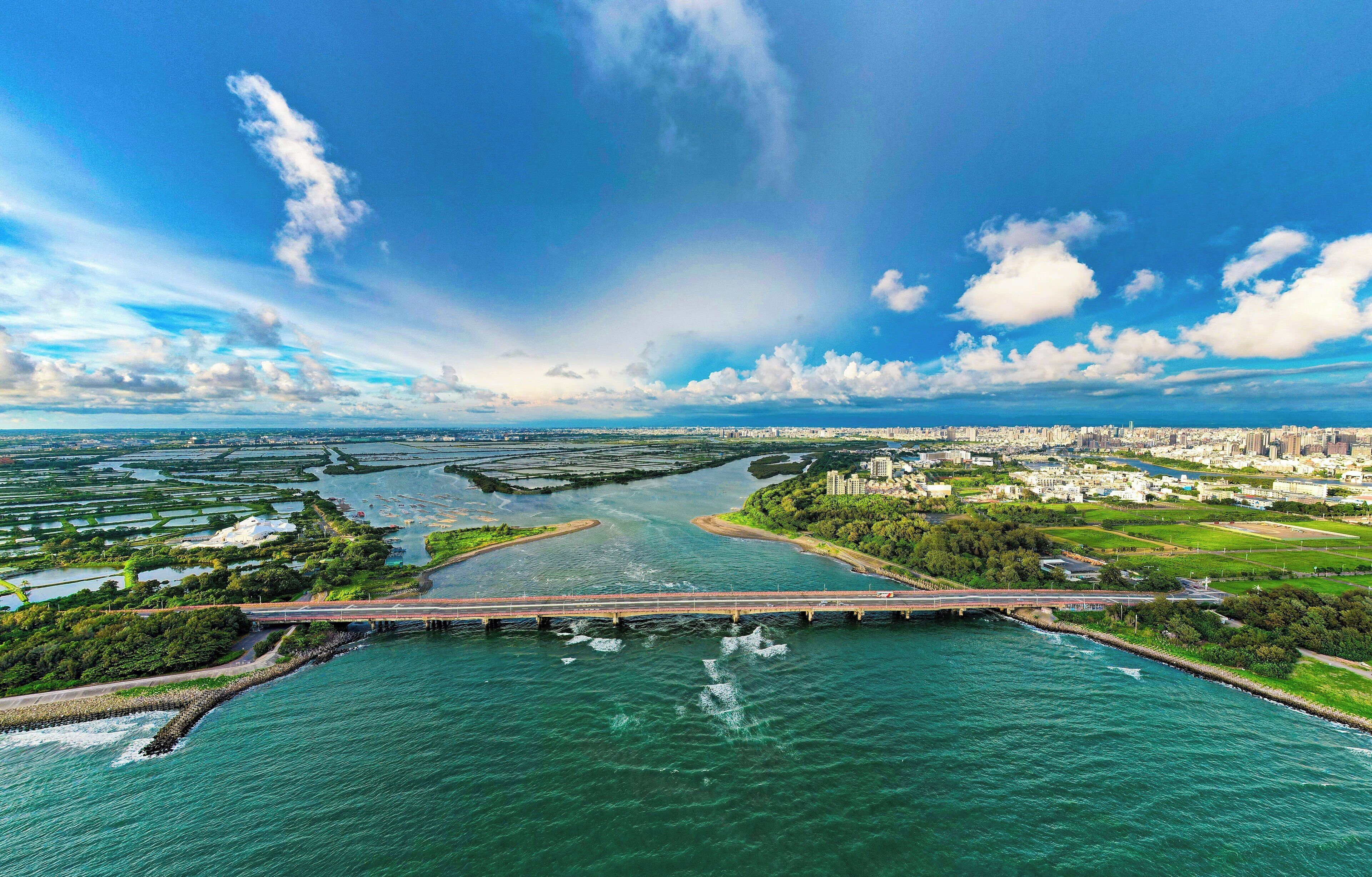 An aerial view of a coastal city with a prominent bridge over water, green areas, and a clear sky.