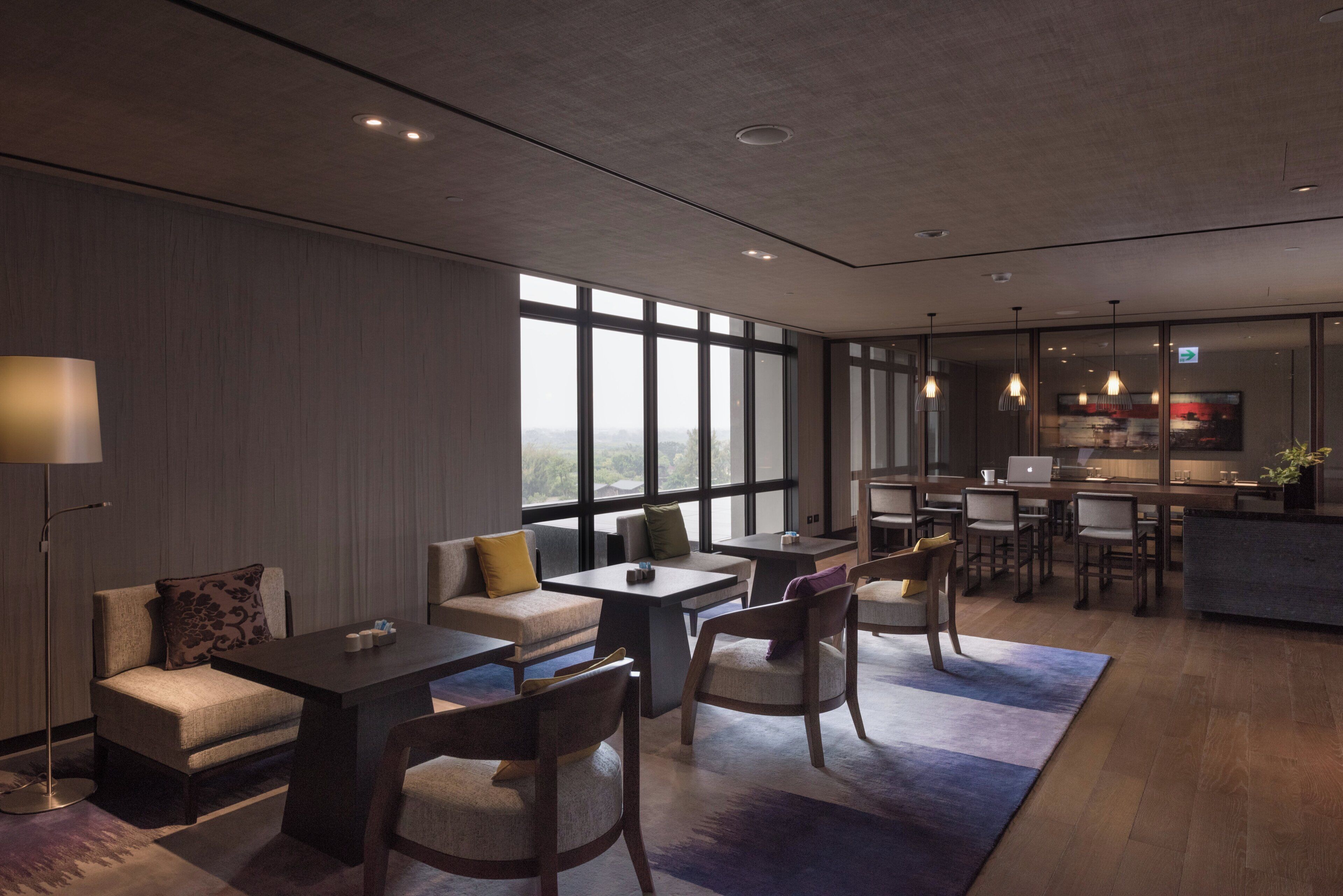 A modern hotel room with a large window, a sofa, a dining table, and a kitchen area.