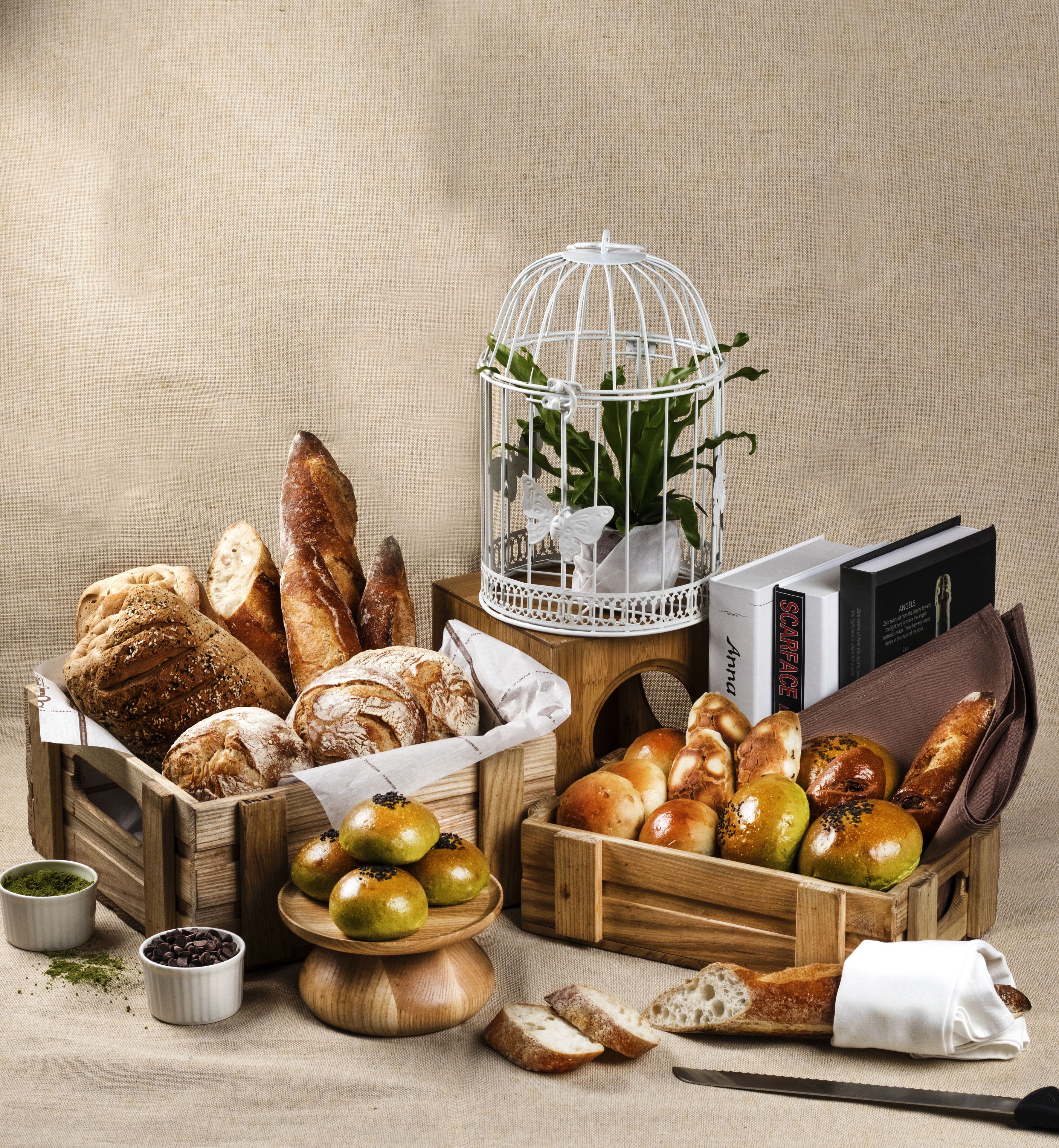 A variety of breads and pastries in wooden crates and baskets, a birdcage with a plant, and a book titled "Scarface".