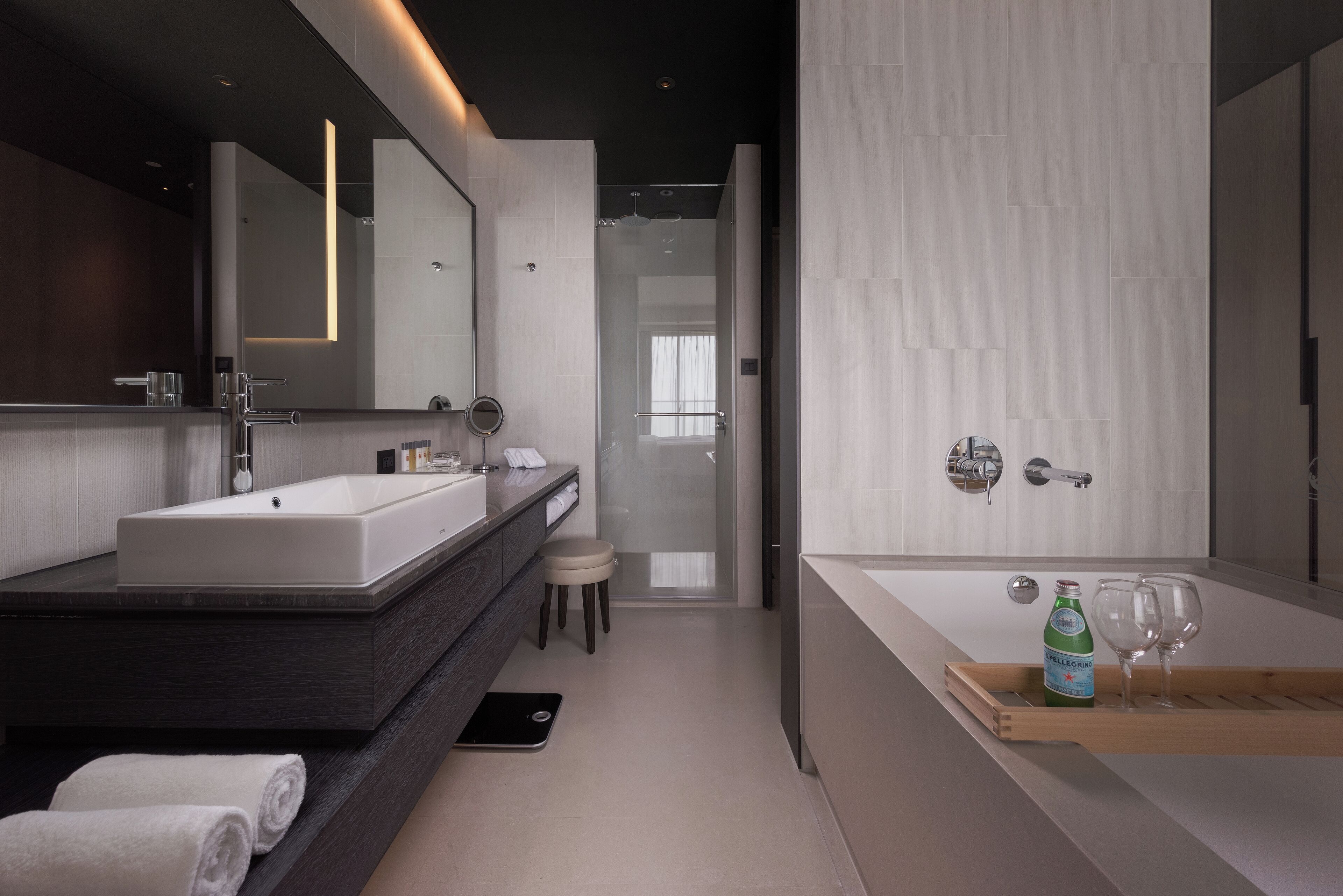 A modern bathroom with a large mirror, a double sink vanity, a shower area, and a wooden tray with a bottle and glasses.