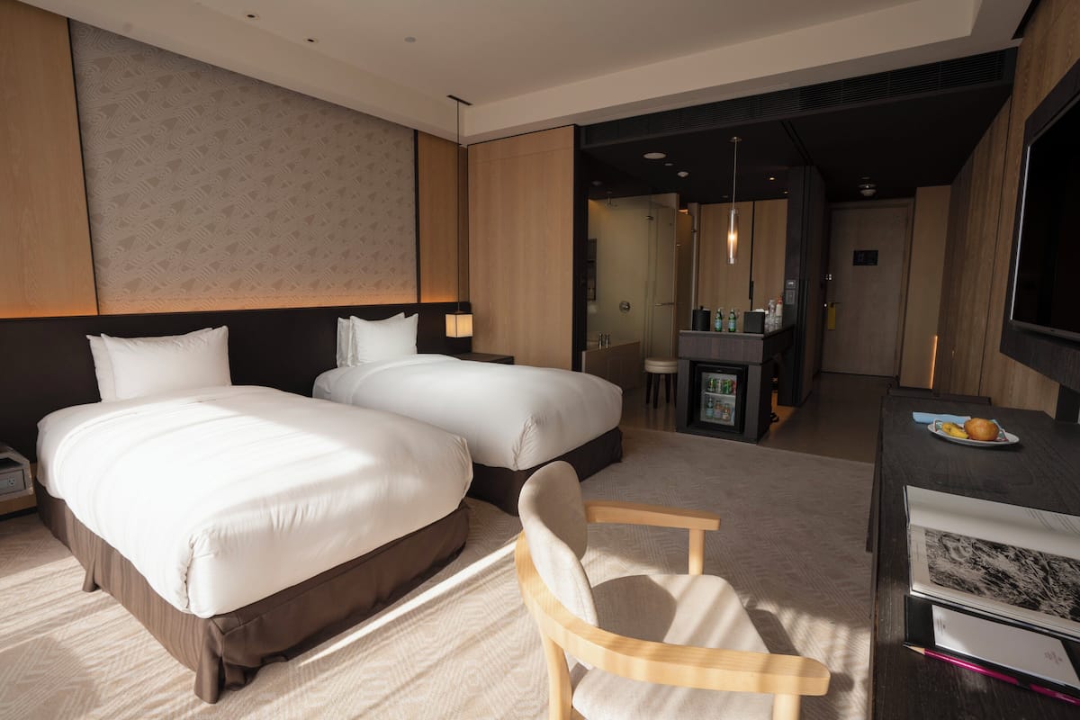 A hotel room with two beds, a desk, a chair, a TV, and a minibar.