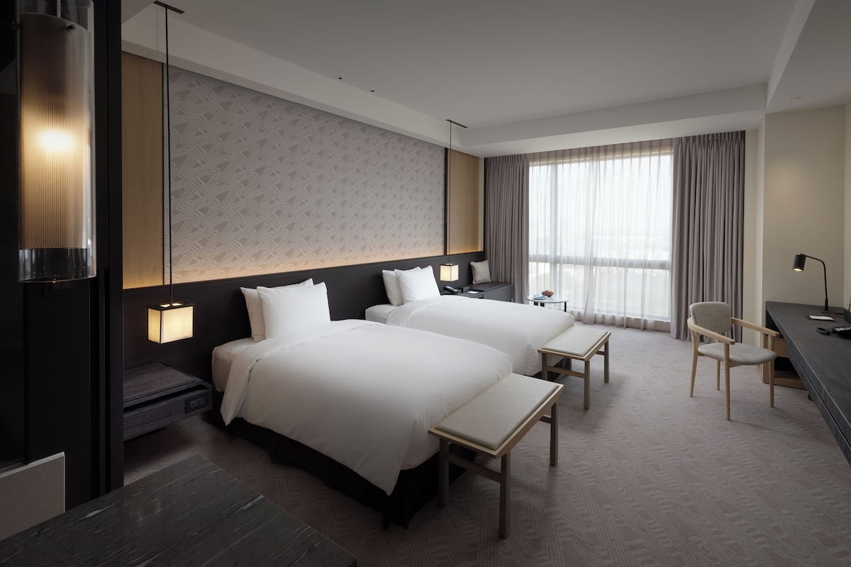 A hotel room with two beds, a desk, a chair, and a large window with curtains.