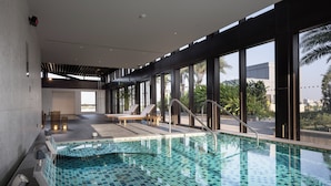 Una piscina interior moderna con baldosas azules cristalinas, tumbonas y grandes ventanales con vistas a palmeras y edificios.