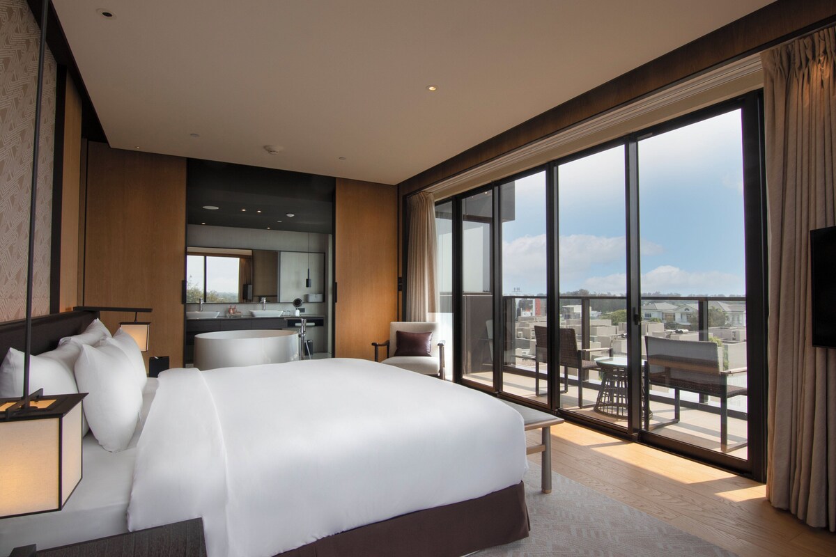 A modern hotel room with a large bed, a bathroom, a balcony with a view, and a flat-screen TV.
