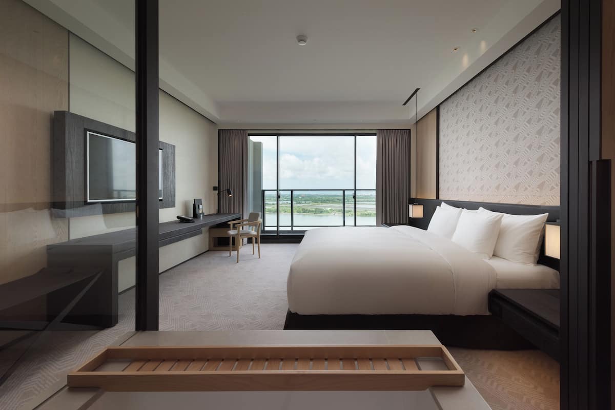 A modern hotel room with a large bed, a desk with a chair, a TV mounted on the wall, and a view of the outdoors.
